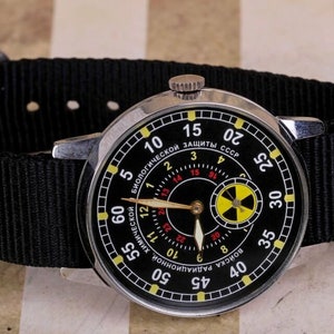 May include: A black wristwatch with a silver band and a black face. The face has a yellow and black radiation symbol and the text "БИОЛОГИЧЕСКОЙ ЗАЩИТЫ СССР" in red.