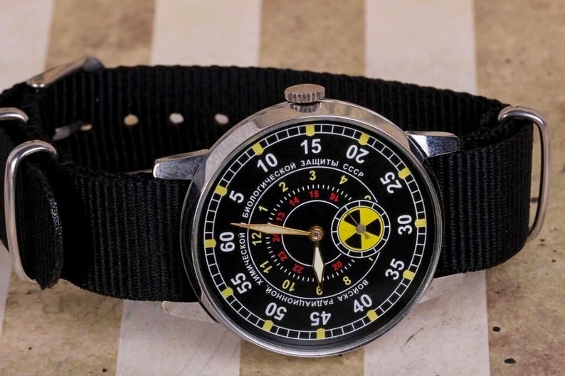 May include: A black wristwatch with a silver band and a black face. The face has a yellow and black radiation symbol and the text "БИОЛОГИЧЕСКОЙ ЗАЩИТЫ СССР" in red.