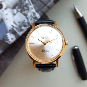 K&ouml;nnte beinhalten: Eine goldfarbene Armbanduhr mit wei&szlig;em Zifferblatt und schwarzem Lederarmband. Das Zifferblatt der Uhr hat schwarze r&ouml;mische Ziffern als Stundenmarkierungen, schwarze Zeiger und den Text "POLJOT de luxe" und "23 jewels shockproof".
