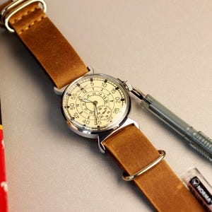 Puede incluir: Un reloj de pulsera de estilo vintage con una esfera blanca y n&uacute;meros negros. El reloj tiene una correa de cuero marr&oacute;n y una caja plateada. El reloj est&aacute; sobre una superficie blanca.