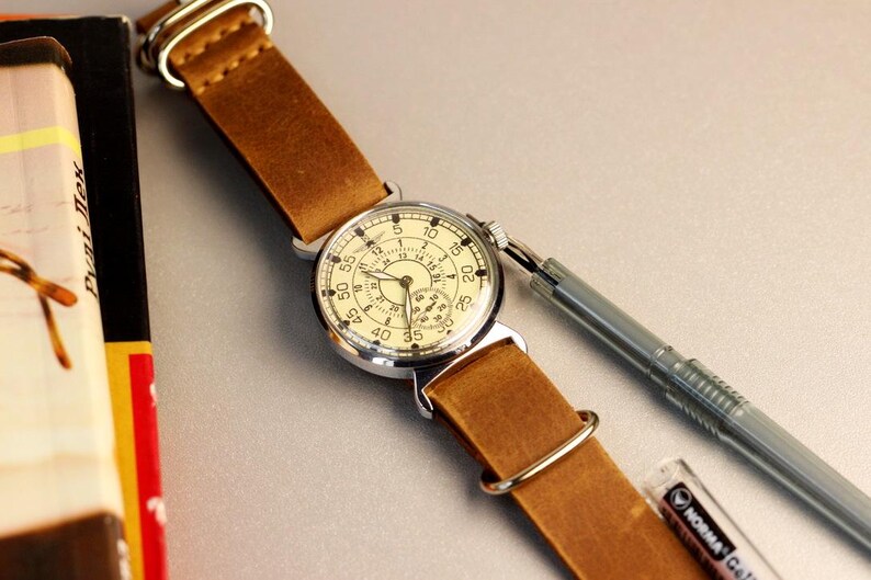 Puede incluir: Un reloj de pulsera de estilo vintage con una esfera blanca y n&uacute;meros negros. El reloj tiene una correa de cuero marr&oacute;n y una caja plateada. El reloj est&aacute; sobre una superficie blanca.