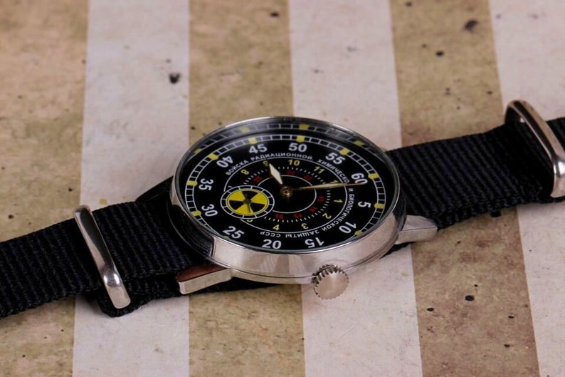 May include: A black wristwatch with a silver case and a black nylon strap. The watch face has a black background with yellow and red markings. The text on the watch face reads "СОЮЗ РАДИАЦИОННОЙ ЗАЩИТЫ СССР".