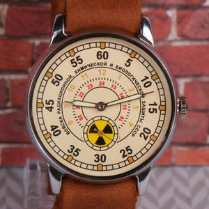 May include: A silver wristwatch with a white face and black hands. The face has a yellow and black radiation symbol in the center and the text "ВОЙСКА РАДИАЦИОННОЙ, ХИМИЧЕСКОЙ И БИОЛОГИЧЕСКОЙ ЗАЩИТЫ СССР" around the outside.