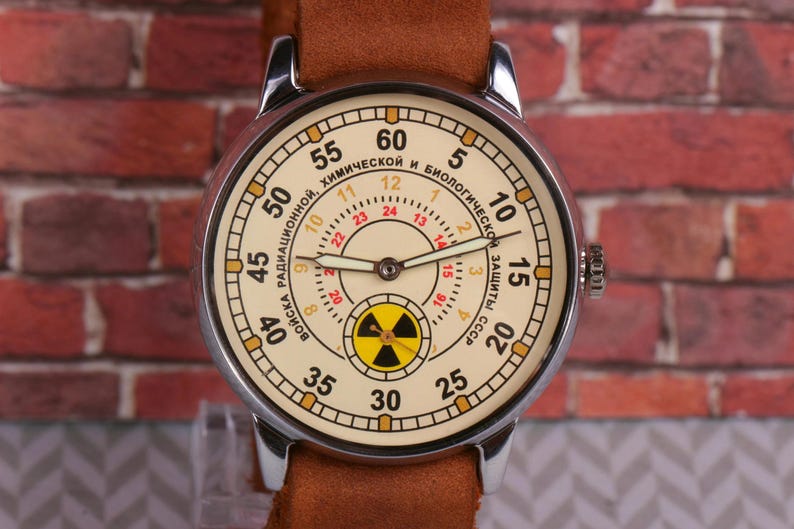 May include: A silver wristwatch with a white face and black hands. The face has a yellow and black radiation symbol in the center and the text "ВОЙСКА РАДИАЦИОННОЙ, ХИМИЧЕСКОЙ И БИОЛОГИЧЕСКОЙ ЗАЩИТЫ СССР" around the outside.
