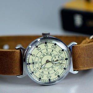 Puede incluir: Un reloj de pulsera plateado con una esfera blanca y una correa de cuero marr&oacute;n. El reloj tiene una esfera de 24 horas y un segundero.