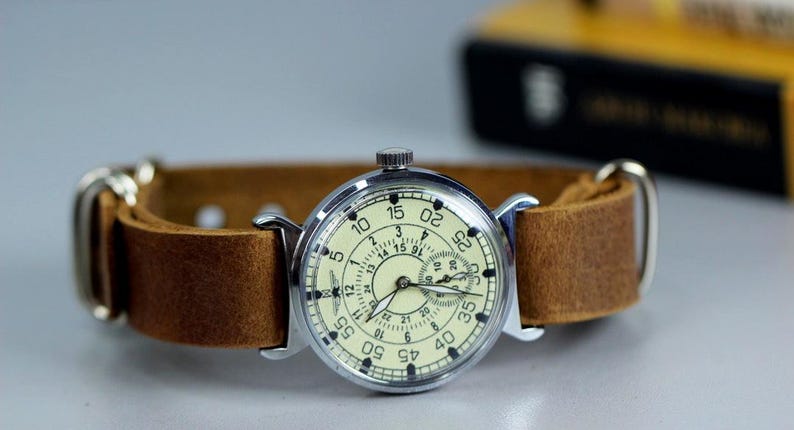 Puede incluir: Un reloj de pulsera plateado con una esfera blanca y una correa de cuero marr&oacute;n. El reloj tiene una esfera de 24 horas y un segundero.