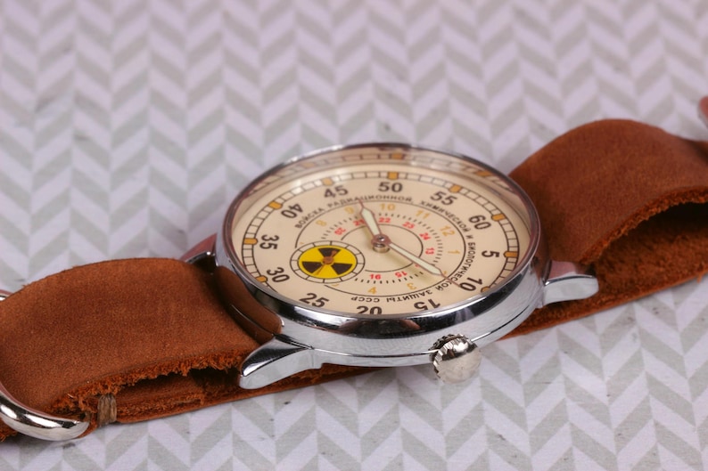 May include: A vintage wristwatch with a brown leather band and a white face with black markings. The watch face has a yellow and black radiation symbol in the center and the text "ЧАСОВЫЙ ЗАВОД ИМ. П.А.  ЧАЙКОВСКОГО СССР" around the edge.