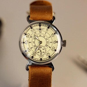 Puede incluir: Un reloj de pulsera vintage con una esfera blanca y marcas negras. El reloj tiene una correa de cuero marr&oacute;n y una caja plateada. La esfera del reloj tiene un reloj de 24 horas y un reloj de 60 segundos.