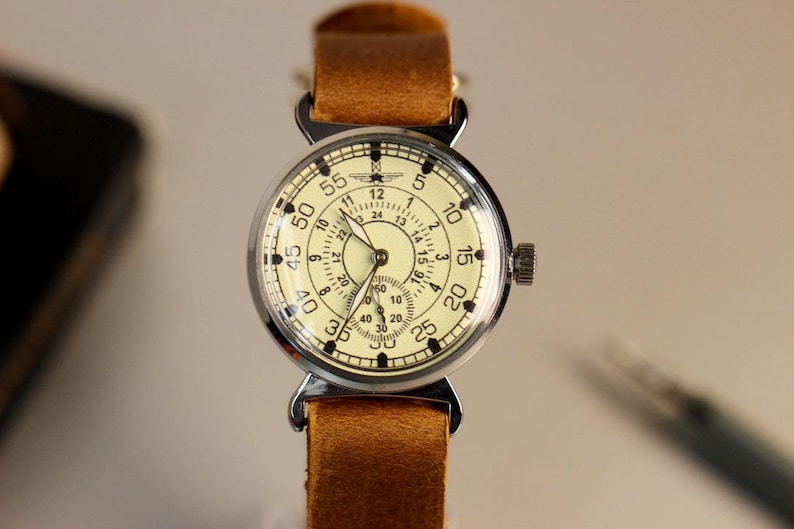Puede incluir: Un reloj de pulsera vintage con una esfera blanca y marcas negras. El reloj tiene una correa de cuero marr&oacute;n y una caja plateada. La esfera del reloj tiene un reloj de 24 horas y un reloj de 60 segundos.