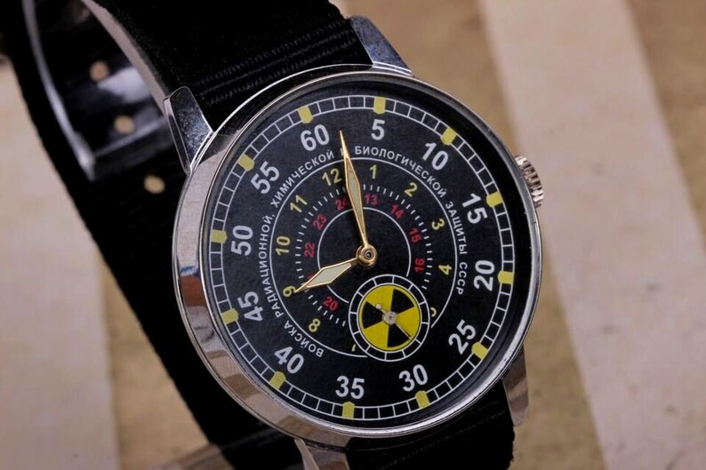 May include: A black wristwatch with a silver case and a black nylon strap. The watch face has a black background with yellow and red markings. The watch face also has a yellow and black radiation symbol in the center. The watch face has the text "РАДИАЦИОННОЙ, ХИМИЧЕСКОЙ" and "ВОИСКА" in Cyrillic script. The watch face also has the text "БИОЛОГИЧЕСКОЙ ЗАЩИТЫ СССР" in Cyrillic script.