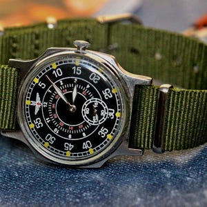 May include: A black and silver wristwatch with a green nylon strap. The watch face has a black background with white numbers and a small second hand dial. The watch has a red airplane symbol on the face.