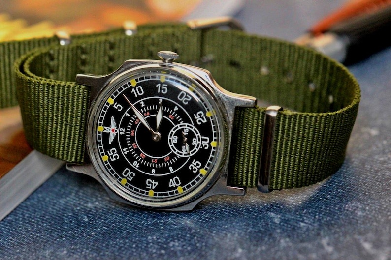 May include: A black and silver wristwatch with a green nylon strap. The watch face has a black background with white numbers and a small second hand dial. The watch has a red airplane symbol on the face.