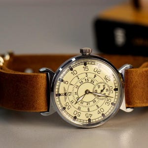Puede incluir: Un reloj de pulsera plateado con una correa de cuero marr&oacute;n. La esfera del reloj tiene un fondo blanco con n&uacute;meros y manecillas negros. El reloj tiene una esfera de 24 horas y un segundero.