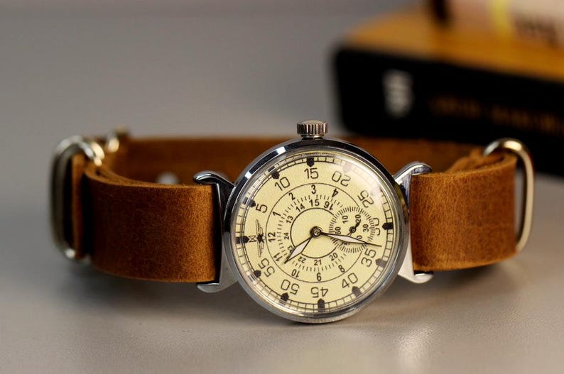 Puede incluir: Un reloj de pulsera plateado con una correa de cuero marr&oacute;n. La esfera del reloj tiene un fondo blanco con n&uacute;meros y manecillas negros. El reloj tiene una esfera de 24 horas y un segundero.