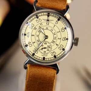 Puede incluir: Un reloj de pulsera vintage con una esfera de color crema y marcas negras. El reloj tiene una caja plateada y una correa de cuero marr&oacute;n.
