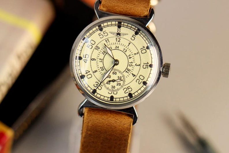 Puede incluir: Un reloj de pulsera vintage con una esfera de color crema y marcas negras. El reloj tiene una caja plateada y una correa de cuero marr&oacute;n.