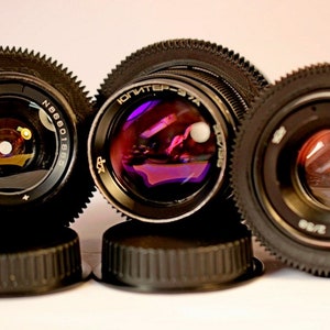 May include: Three vintage camera lenses with black metal bodies and silver accents. The lenses have a black and silver gear ring around the edge. The lenses are sitting on black lens caps.