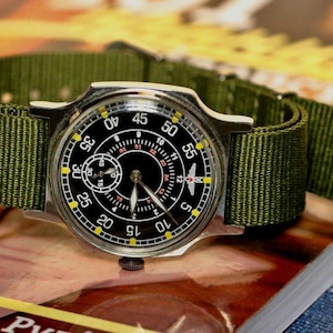 May include: A black and silver wristwatch with a green nylon strap. The watch face has a black background with white markings and a small subdial. The watch has a yellow outer ring with black numbers.