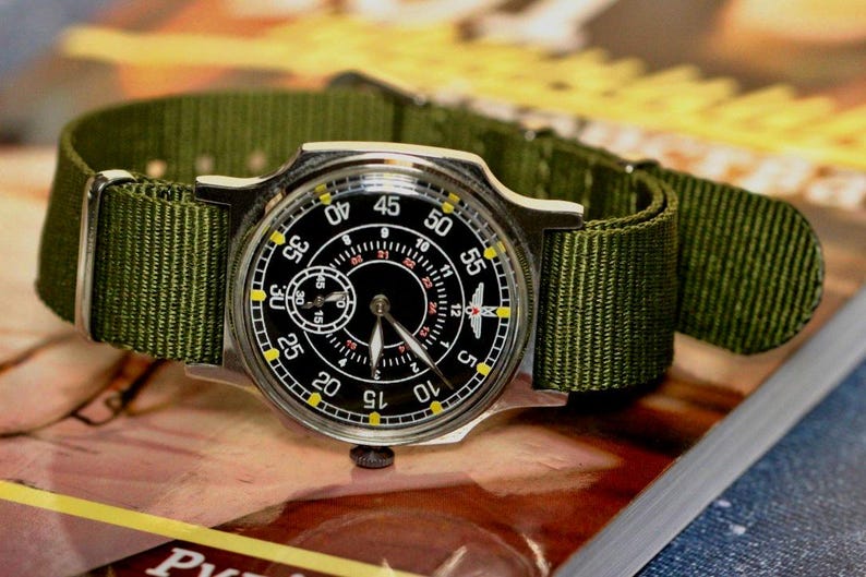 May include: A black and silver wristwatch with a green nylon strap. The watch face has a black background with white markings and a small subdial. The watch has a yellow outer ring with black numbers.