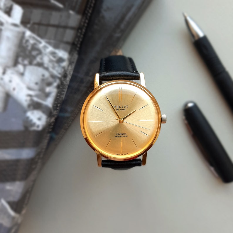 May include: A gold-tone wristwatch with a black leather strap. The watch face is gold with black markings and the text "Poljot De Luxe 23 Jewels Shockproof Made in USSR".