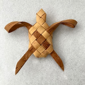 May include: A woven turtle made from natural materials, with a light brown body and darker brown legs and head. The turtle is crafted with a traditional weaving technique, creating a unique and intricate design.