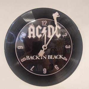 Könnte beinhalten: Schwarze Wanduhr mit dem AC/DC-Logo und der Aufschrift "BACK IN BLACK". Das Zifferblatt der Uhr ist als Schallplatte gestaltet. Die Zeiger der Uhr sind weiß.