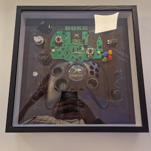 Könnte beinhalten: Gerahmte Präsentation eines Xbox Duke-Controllers und einer Leiterplatte. Der Controller ist schwarz mit einem grünen Xbox-Logo. Die Leiterplatte ist grün mit dem Wort "DUKE" oben. Der Rahmen ist schwarz.