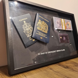 Op de afbeelding: Een zwarte lijst met een schaduwdoos met daarin een Canadees paspoort, een Brits paspoort, een stapel biljetten en een creditcard. De tekst "IN GEVAL VAN NOOD GLAS BREKEN" staat onderaan de lijst.