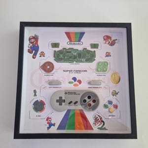 Peut inclure: Œuvre encadrée présentant un contrôleur Super Famicom démonté et ses composants. Le design comprend un arc-en-ciel, des personnages de Mario et des étiquettes textuelles. Le cadre est noir et l'œuvre est affichée sur un fond blanc.