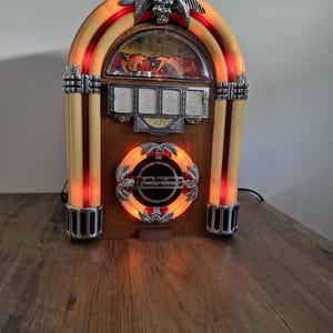 May include: A tabletop jukebox featuring a wooden body with an arched top, illuminated by internal red and orange lights. It includes silver palm tree accents, a central circular speaker grille with the text Jukebox, and a selection panel display.