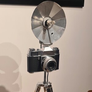 Peut inclure: Un appareil photo vintage argenté avec un corps noir et un flash monté sur un trépied en bois.