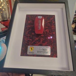 May include: Framed Ferrari key in a black frame. The red key is displayed on a textured red background. A silver plaque below reads "Ferrari MODENA ITALY Special Edition."