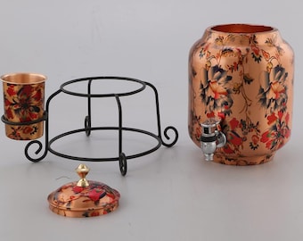 Könnte beinhalten: Kupferner Wasserdispenser mit Blumenmuster, einer passenden Tasse und einem Ständer. Der Dispenser hat einen Hahn und einen Deckel mit einem Knopf.