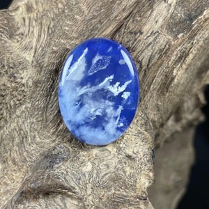 May include: An oval-shaped, polished gemstone with a deep blue base and white, cloud-like inclusions. The stone has a glossy finish and is set against a textured, brown wooden surface. The gemstone's design resembles a cloudy sky.