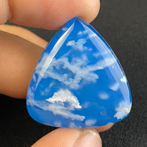 Puede incluir: Un cabujón de ágata azul triangular con patrones blancos en forma de nubes. La ágata es translúcida y tiene un acabado brillante.