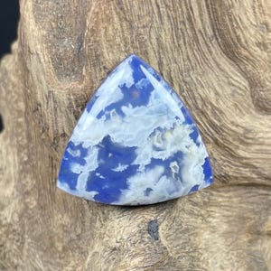 May include: A triangular gemstone with a blue and white cloud-like pattern. The polished stone has a glossy finish and is set against a textured, brown wooden surface. The gemstone's shape is a triangle.