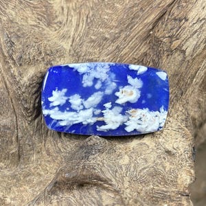 May include: A rectangular, polished gemstone with a vibrant blue base, resembling a daytime sky. White, cloud-like patterns are dispersed throughout the stone. The gemstone is set against a textured, wooden surface.