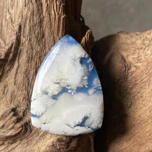 Könnte beinhalten: Ein polierter, dreieckiger Edelstein-Cabochon mit einer durchscheinenden blauen Basis und undurchsichtigen, wolkenartigen weißen dendritischen Einschlüssen, die ein geschichtetes, atmosphärisches Erscheinungsbild auf der glatten Oberfläche erzeugen.