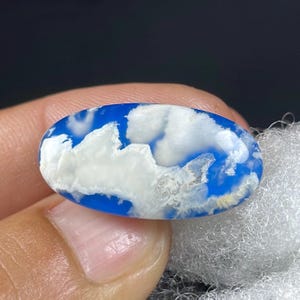 May include: An oval gemstone with a bright blue background and white, cloud-like patterns. The polished stone is held between fingers. The gemstone's design evokes a cloudy sky. The stone's measurements are in centimetres.