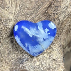 May include: A heart-shaped gemstone with a blue and white cloud-like pattern. The polished stone has a glossy finish and is set against a textured, wooden background. The heart shape is clearly defined.