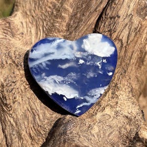 May include: A heart-shaped stone with a blue and white cloud-like pattern. The stone is polished and smooth, with a deep blue base color and white swirls. The heart is displayed on a textured, brown wooden surface.