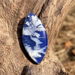May include: A polished, elongated gemstone with a deep blue base and white, cloud-like patterns. The stone is set against a natural wood background, highlighting its unique design and color variations. The gemstone's shape is reminiscent of a leaf or an eye.