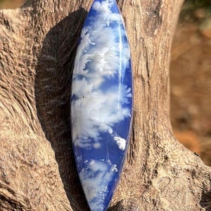 May include: A blue and white pendant with a cloud-like design. The elongated, teardrop-shaped pendant features a glossy finish and is set against a natural wood background. The blue resin is swirled with white, creating a unique pattern.