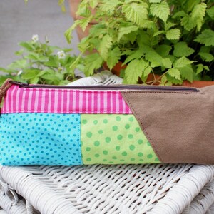 Könnte beinhalten: Eine farbenfrohe Patchwork-Reißverschlusstasche mit rosa-weißen Streifen, blau-weißen Punkten, grün-weißen Punkten und braunem Stoff. Die Tasche liegt auf einer weißen Weidenoberfläche.