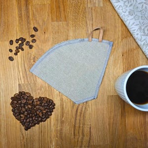 Könnte beinhalten: Ein hellblauer Kaffeefilter mit Ledergriff, Kaffeebohnen in Herzform und eine Tasse Kaffee auf einer Holzoberfläche. Ein Stoff mit Blumenmuster ist ebenfalls vorhanden.