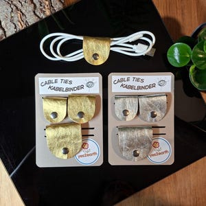 Pode incluir: Laços de cabo em couro dourado e prateado com fechos de pressão, exibidos em cartões com o texto "Cable Ties Kabelbinder". Um cabo de carregamento branco é preso com um laço de cabo de couro dourado. Da yes2earth.