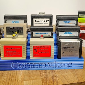 Può includere: Una collezione di cartucce di giochi e accessori Commodore 64 vintage, tra cui "Donkey Kong" e "Turbo232", esposta su un supporto blu con il logo Commodore. Le cartucce sono di vari colori e presentano etichette di testo.