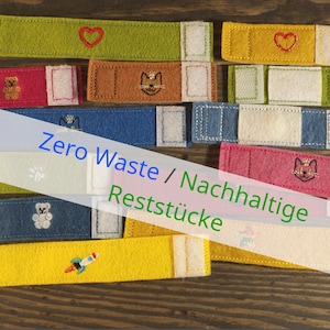 Könnte beinhalten: Eine Auswahl an farbigen Filzstreifen mit gestickten Motiven wie Herzen, Katzen und einer Rakete. Der Text "Zero Waste / Nachhaltige Reststücke" ist überlagert. Jeder Streifen hat einen Klettverschluss.