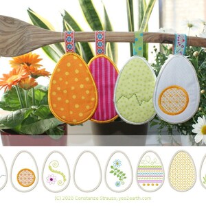 Puede incluir: Cinco adornos de huevos de Pascua bordados con diferentes diseños, que incluyen lunares, rayas, un huevo roto, una yema y una flor. Los adornos están colgados de una cuchara de madera.