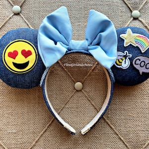 May include: Blue denim Minnie Mouse ears with a light blue bow. The ears are decorated with patches including a yellow smiley face with red hearts for eyes, a bee, a star with a rainbow, and a purple speech bubble with the word "cool".